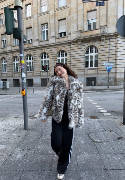 Mujer joven con abrigo de piel con manchas y diadema caminando por la acera de la ciudad cerca de una intersección y un edificio de piedra con ventanas arqueadas.