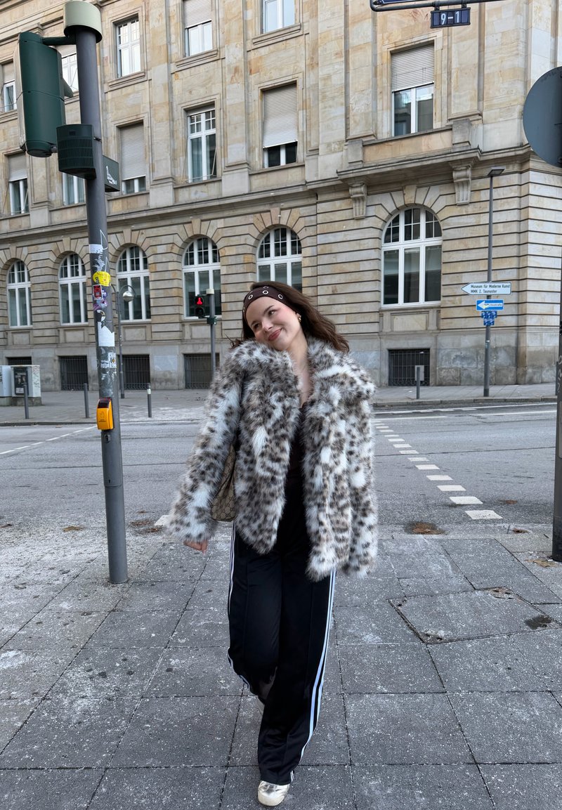 Jeune femme portant un manteau en fourrure à pois et un bandeau, marchant sur le trottoir en ville près d'une intersection et d'un bâtiment en pierre avec des fenêtres en arc.