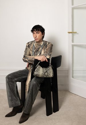 Persona vestida con un conjunto y botas con estampado de piel de serpiente, sentada en una silla negra, sosteniendo un bolso a juego con el mismo estampado en una habitación interior luminosa.