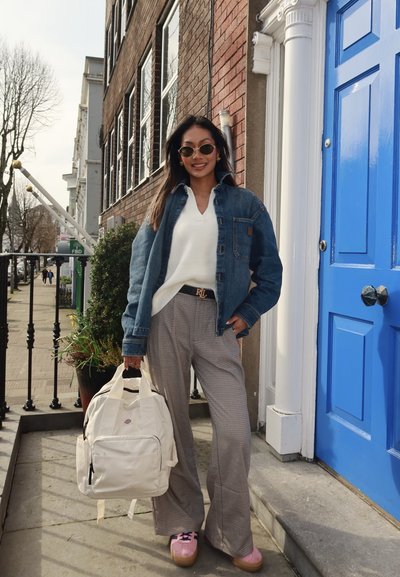 Chaqueta de mezclilla, suéter de punto blanco, pantalones de cuadros, llevando una mochila color crema, con zapatos de plataforma rosa, frente a una puerta azul en el fondo.