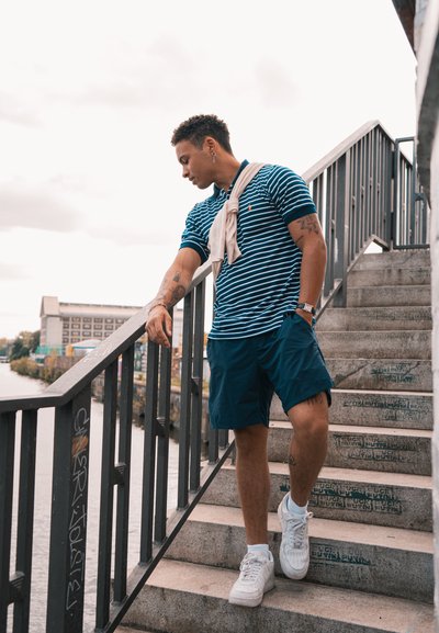 Camiseta polo a rayas azules, pantalones cortos navy y zapatillas blancas. De pie en unas escaleras con un pasamanos, mirando hacia un canal. Brazo tatuado visible.