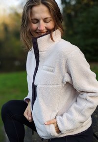 Casaco de fleece bege claro com colarinho alto, detalhes pretos e um logotipo. Apresenta um fecho éclair à frente e bolsos laterais.