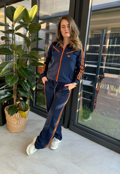 Chándal de color marino con rayas naranjas, que incluye una chaqueta con cremallera y pantalones a juego. Zapatillas blancas que complementan el aspecto deportivo. Entorno interior con plantas.