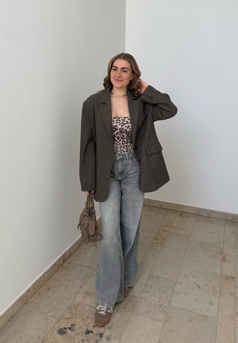 Giovane donna sorridente, indossa un blazer scuro oversize, top con stampa leopardata, jeans a gamba larga, sneakers, tiene in mano una borsa taupe, in piedi in una stanza dai toni neutri.