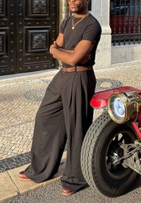 Camicia nera a maniche corte abbinata a pantaloni neri larghi. Cintura marrone con finitura testurizzata. Scarpe marrone con zeppa. Veicolo rosso con fari rotondi.