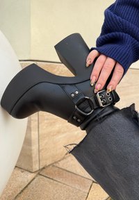 Čierna platformová čižma s zaobleným dizajnom, vybavená prackovým remienkom a striebornými kovovými prvkami. Textúrovaný materiál a robustná podrážka. Nosí sa s roztrhanými džínsami.