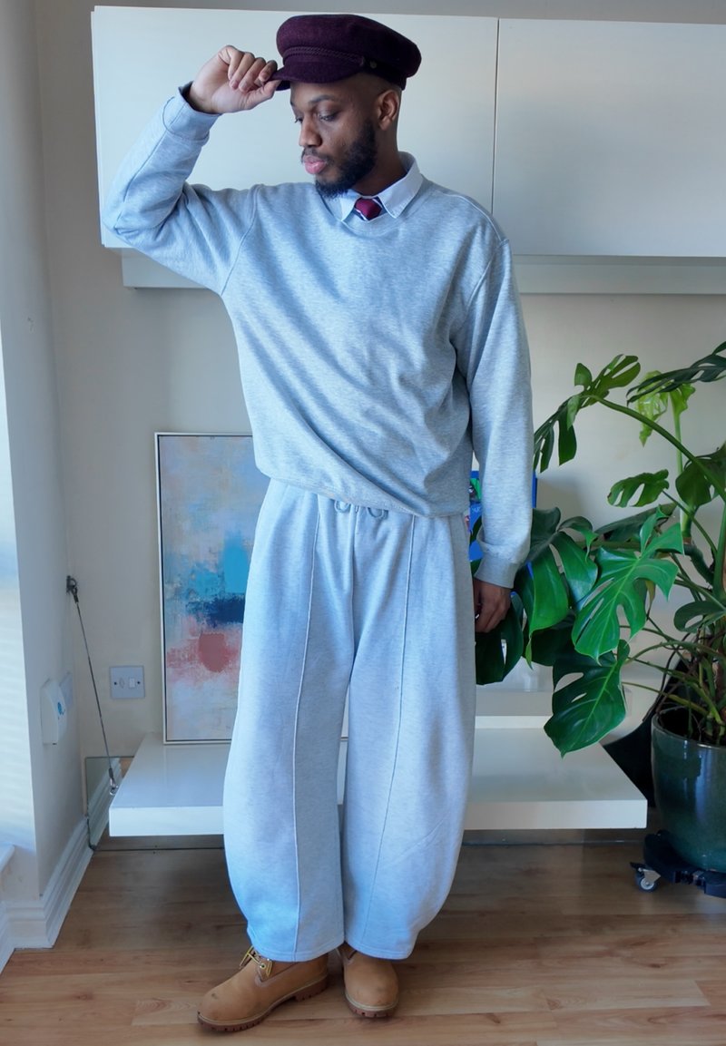 Homme portant un sweat-shirt gris et un pantalon de survêtement large, une casquette marron et des bottes, debout à l'intérieur près d'une peinture abstraite et d'une grande plante verte.