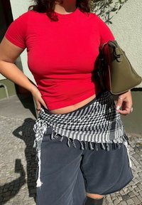 Punane keha lähedal t-särk, mustad lühikesed püksid ja must-valge mustriga sall, mis on ümber vöö seotud. Oliivroheline käekott. Päikseline välistingimus.