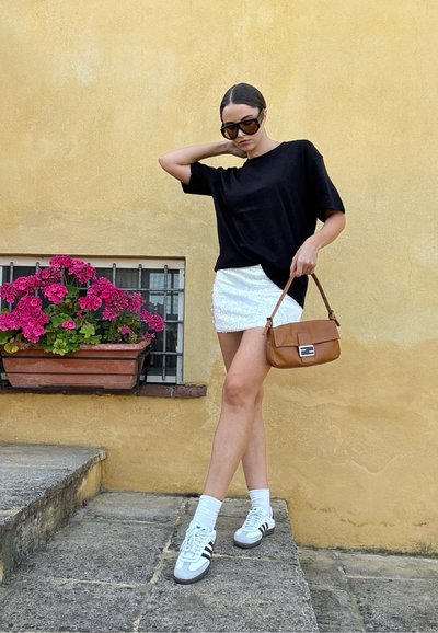 Camiseta negra de gran tamaño, falda mini blanca brillante, bolso cruzado marrón con cierre, calcetines blancos, zapatillas gris y negras. Flores rosas en el fondo.