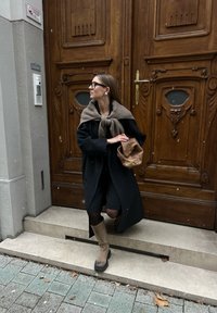 Μαύρο παλτό και καφέ κασκόλ πάνω από ένα σκούρο ντύσιμο, συνδυασμένα με ψηλές καφέ μπότες. Κρατώντας μια καφέ τσάντα, στέκεται δίπλα σε μια ξύλινη πόρτα με περίτεχνες λεπτομέρειες.