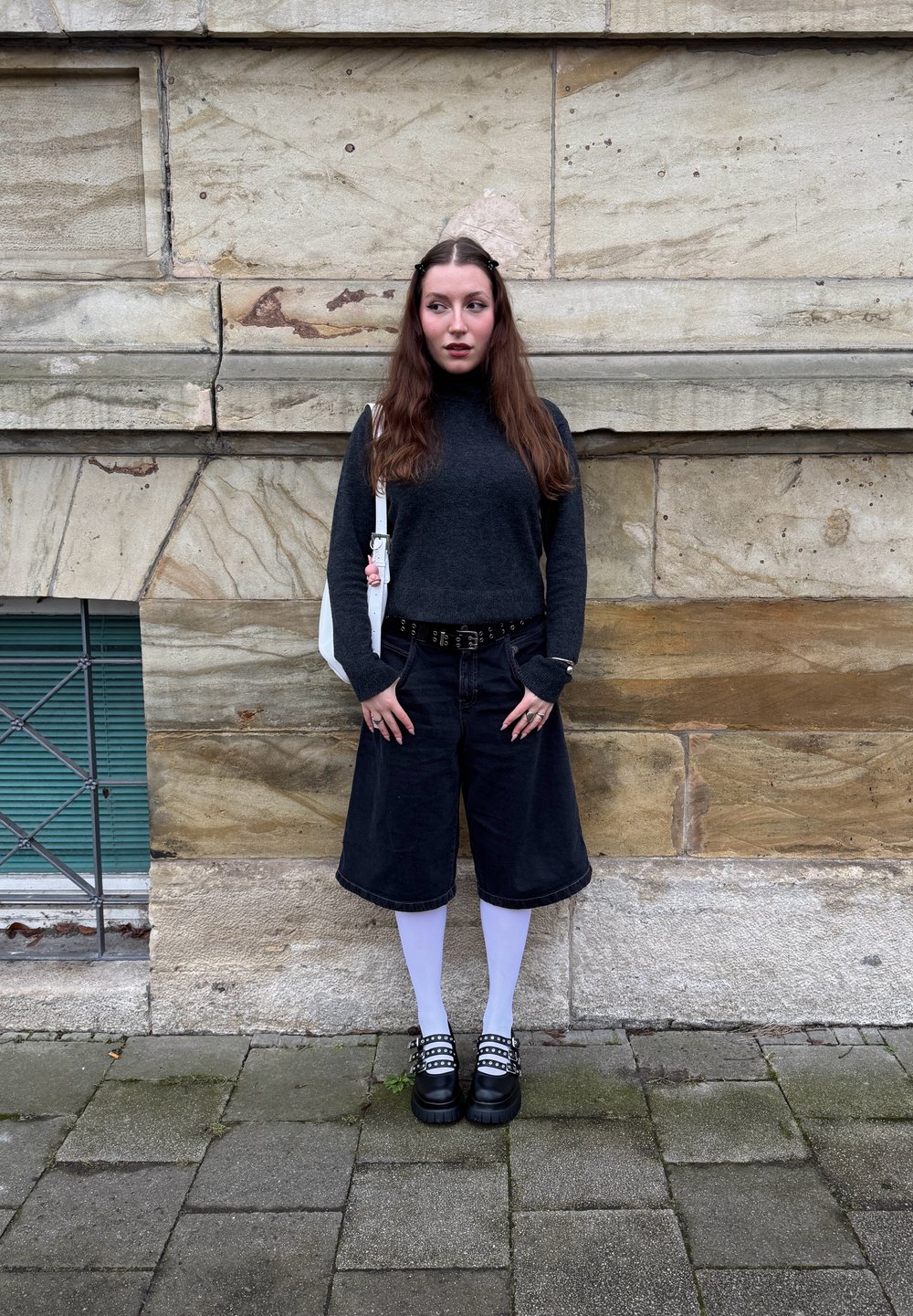 Pessoa a usar um suéter gola alta cinza escuro, calças culottes de denim preto, meias brancas até a coxa e sapatos plataforma pretos chunky, com uma bolsa branca.