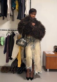 Veste en fausse fourrure marron, jupe en jean déchirée avec fente haute, sac à main noir brillant avec des accents argentés, et escarpins noirs à bout pointu.