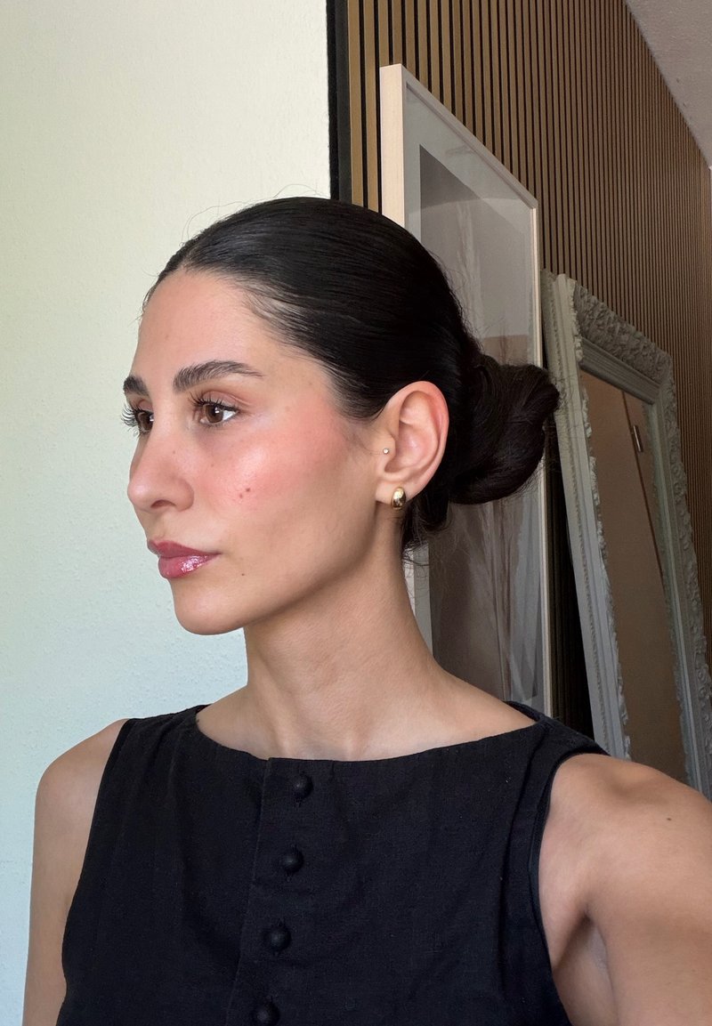 Femme aux cheveux noirs attachés en chignon et portant de petites boucles d'oreilles en or, vêtue d'un haut noir sans manches, debout à l'intérieur près d'œuvres d'art encadrées et d'un miroir.