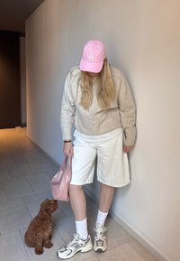 Eine Person trägt einen beigen Pullover und weiße Shorts, während sie eine rosa Tasche hält. Ein kleiner brauner Hund steht neben ihrem Bein. Im Hintergrund sind helle Fliesen und eine schlichte Wand.