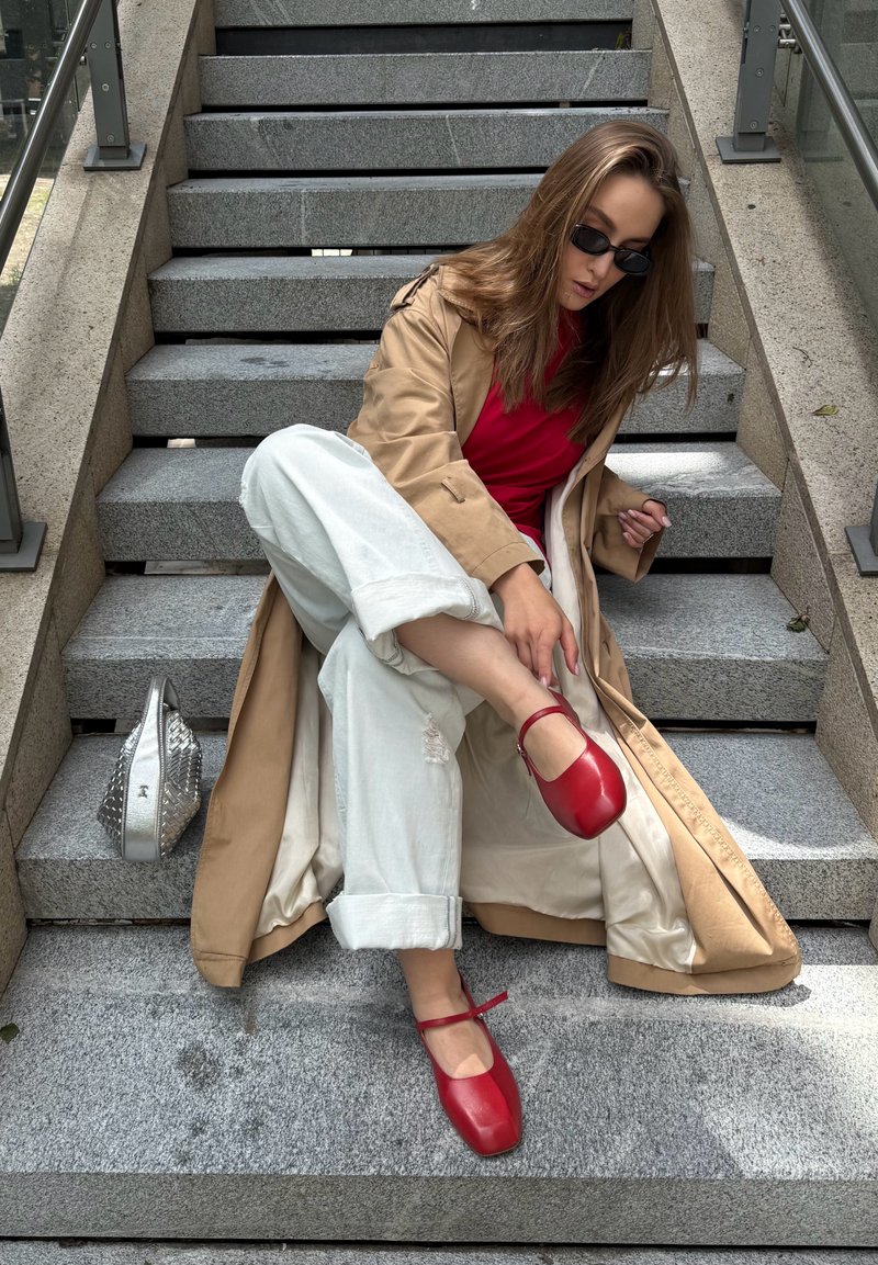 Femme en trench beige, haut rouge, pantalon blanc et chaussures rouges assise sur des escaliers en pierre à l'extérieur, ajustant sa chaussure avec un sac à main argenté à côté d'elle.