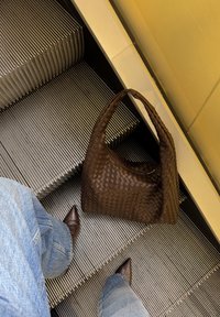 Ruskea punottu nahkainen käsilaukku pehmeällä hobo-muodolla, joka lepää metallisten liukuportaiden askelmilla pisteellisten ruskeiden kenkien ja farkkujen vieressä.