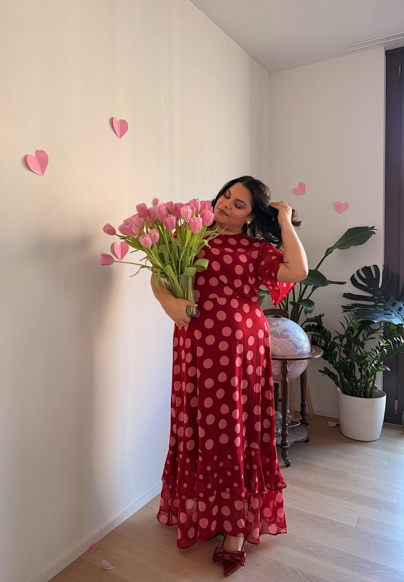 Femme en robe rouge à pois tenant des tulipes roses, debout près de décorations en forme de cœur et de plantes d'intérieur dans une pièce lumineuse avec un sol en bois.