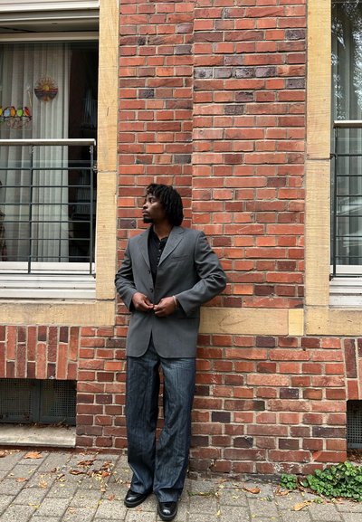 Blazer gris sobre una camisa negra, combinado con jeans de mezclilla azul oscuro y zapatos negros. De pie contra una pared de ladrillos rojos con acentos beige.