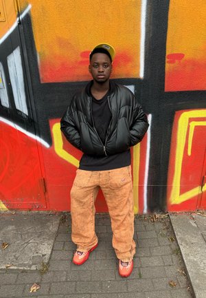 Joven vestido con una chaqueta negra, gorra amarilla, pantalones naranjas y zapatos rojos, de pie con las manos en los bolsillos frente a una pared de graffiti colorido.