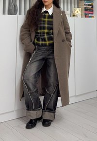 Manteau en laine marron par-dessus un pull à carreaux noir et jaune, associé à un jean foncé déchiré et des chaussures noires. Col blanc visible.