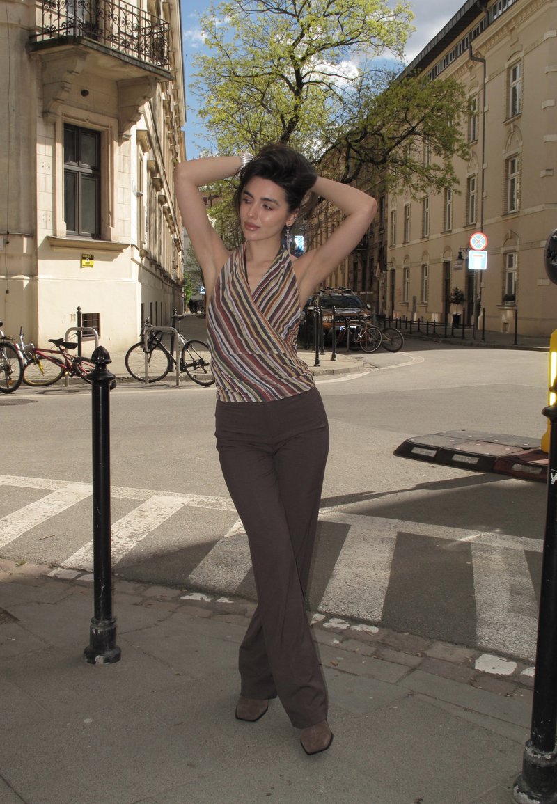Femme vêtue d'un haut sans manches rayé et d'un pantalon sombre, debout dans une rue de la ville, posant les mains derrière la tête près de vélos et de bâtiments.