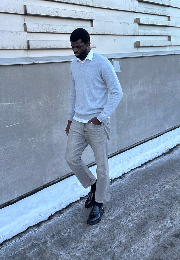 Moški v sivi jopi, beli srajci, svetlih kavbojkah in črnih čevljih hodi po zasneženi pločniku ob betonskem zidu.