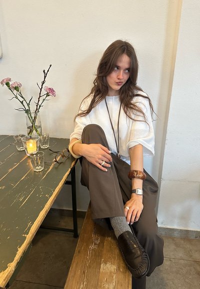Joven con el pelo largo sentada en un banco de madera, vestida con una blusa blanca, pantalones marrones, zapatos tipo mocasín y accesorios, junto a una mesa con flores y una vela.