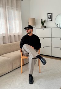 Pull noir côtelé, pantalon gris, chaussures noires, assis sur une chaise en bois sur un tapis clair, avec une lampe moderne et une plante en arrière-plan.