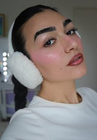 Weiße, flauschige Ohrenschützer bedecken die Ohren und passen zu einem hellen Shirt. Das Gesicht ist mit zartem, rosa Rouge und glänzenden Lippen betont.
