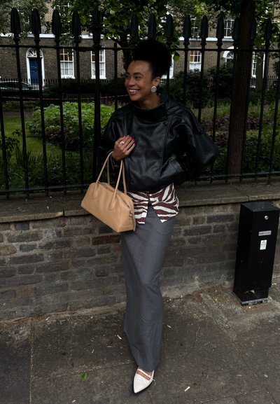 Mujer con chaqueta de cuero negra y camisa con estampado de cebra sosteniendo un bolso beige, de pie en la acera junto a una valla de hierro y una pared de ladrillos, sonriendo.