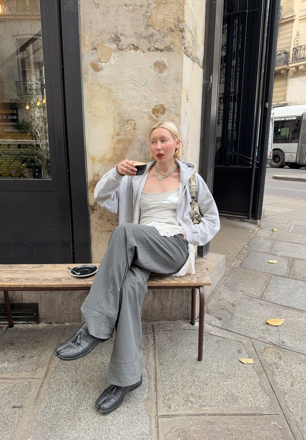 Grå oversized hettegenser, hvit singlet med blonder, grå vidbukser og teksturerte svarte sko. Sitter på en trebenk med kaffe.