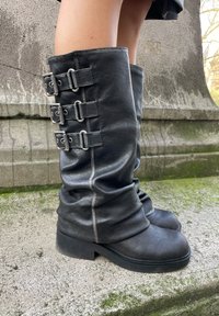 Črne usnjene visokodevske škornje s štirimi nastavljivimi kovinskimi zaponkami na strani, mehak material, okrogla konica in nizka blok peta.