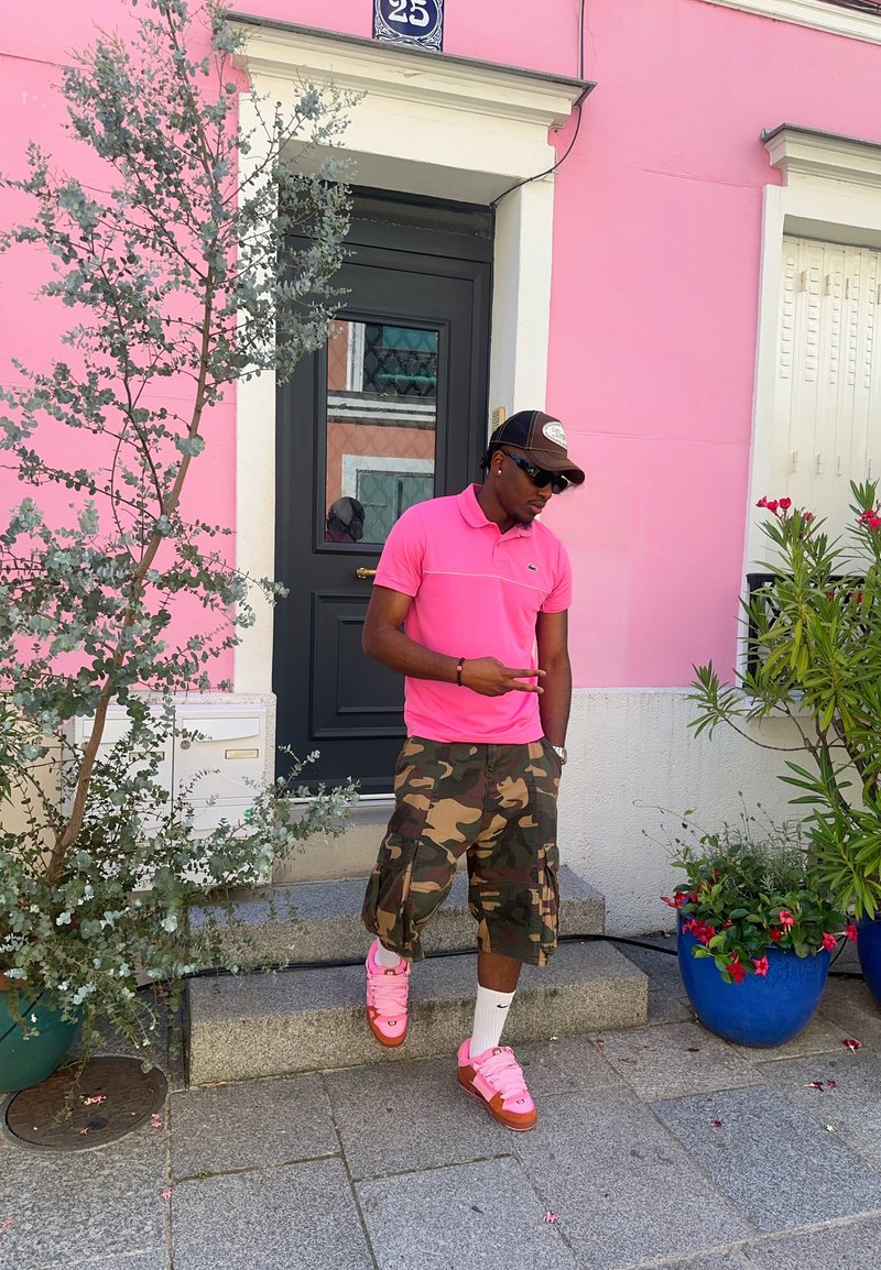 Homme en chemise rose et short camouflage posant avec le signe de la paix devant un bâtiment rose avec une porte noire et des plantes en pot.