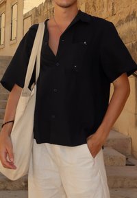Zwarte kortemouwen shirt met een borstzak, met knoopsluiting. Draagt met lichte linnen broek en een canvas totebag.