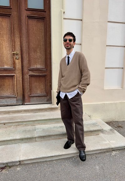 Jersey de punto beige sobre una camisa blanca, corbata negra, pantalones marrones y zapatos negros. De pie en unos escalones junto a una puerta de madera con paneles.