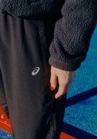 Crne sportske hlače s bijelim Asics logom na bedru, uparene s tamnom mekanom jaknom. Ruka s crvenim lakom za nokte odmara se sa strane.