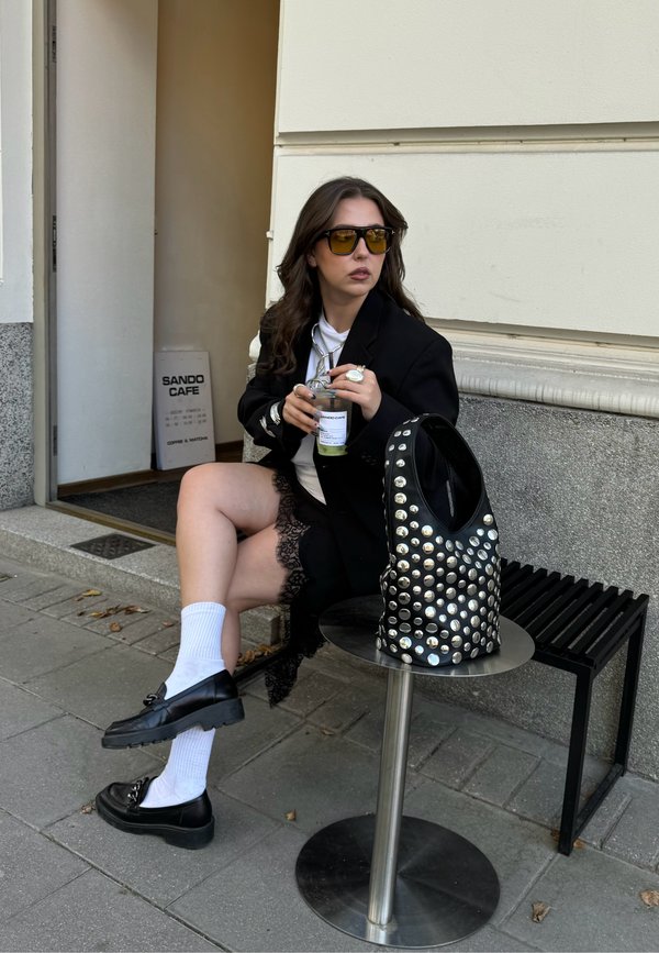 Sac en cuir noir à clous, blazer noir surdimensionné, chemise blanche, jupe à volants en dentelle, chaussures noires à plateforme et chaussettes blanches. Ambiance de café en plein air.