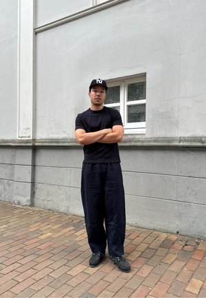 Camiseta negra de manga corta combinada con pantalones oscuros de corte holgado y zapatos negros. La persona lleva una gorra negra con un logo.