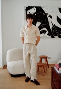 Camisa de botão de manga curta, bege, com textura ribada, combinada com calças largas de um tom bege claro e sapatos mocassim pretos, em um ambiente moderno interior.