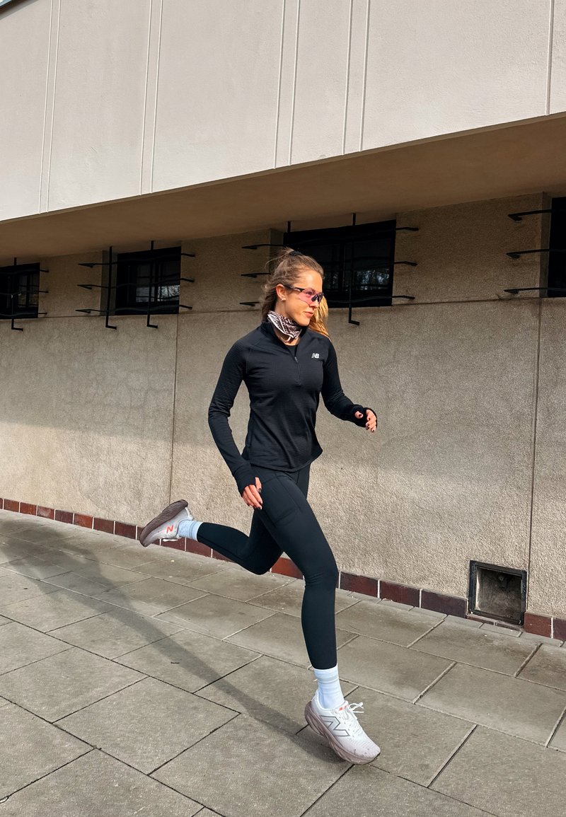 Žena v černém sportovním oblečení a bílých teniskách běží po chodníku vedle béžové budovy s malými mřížkovanými okny.