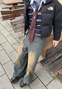 Chaqueta acolchada negra sobre una camisa de rayas y una corbata de cuadros; jeans anchos, desgastados y grises; zapatos de plataforma negros y brillantes.