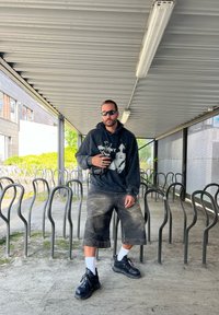 Felpa nera oversize con stampa grafica, pantaloni shorts grigi, sneakers neri e calzini bianchi, in piedi in un'area coperta con rastrelliere per biciclette.