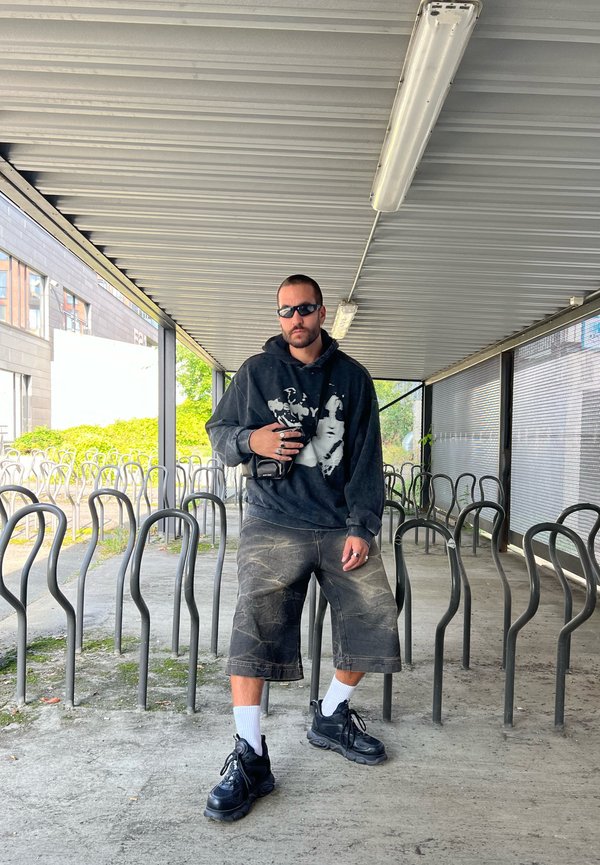Čierna oversized mikina s grafickým potlačou, šedé šortky, čierne tenisky a biele ponožky, stojí v prístrešku s bicyklovými stojanmi.