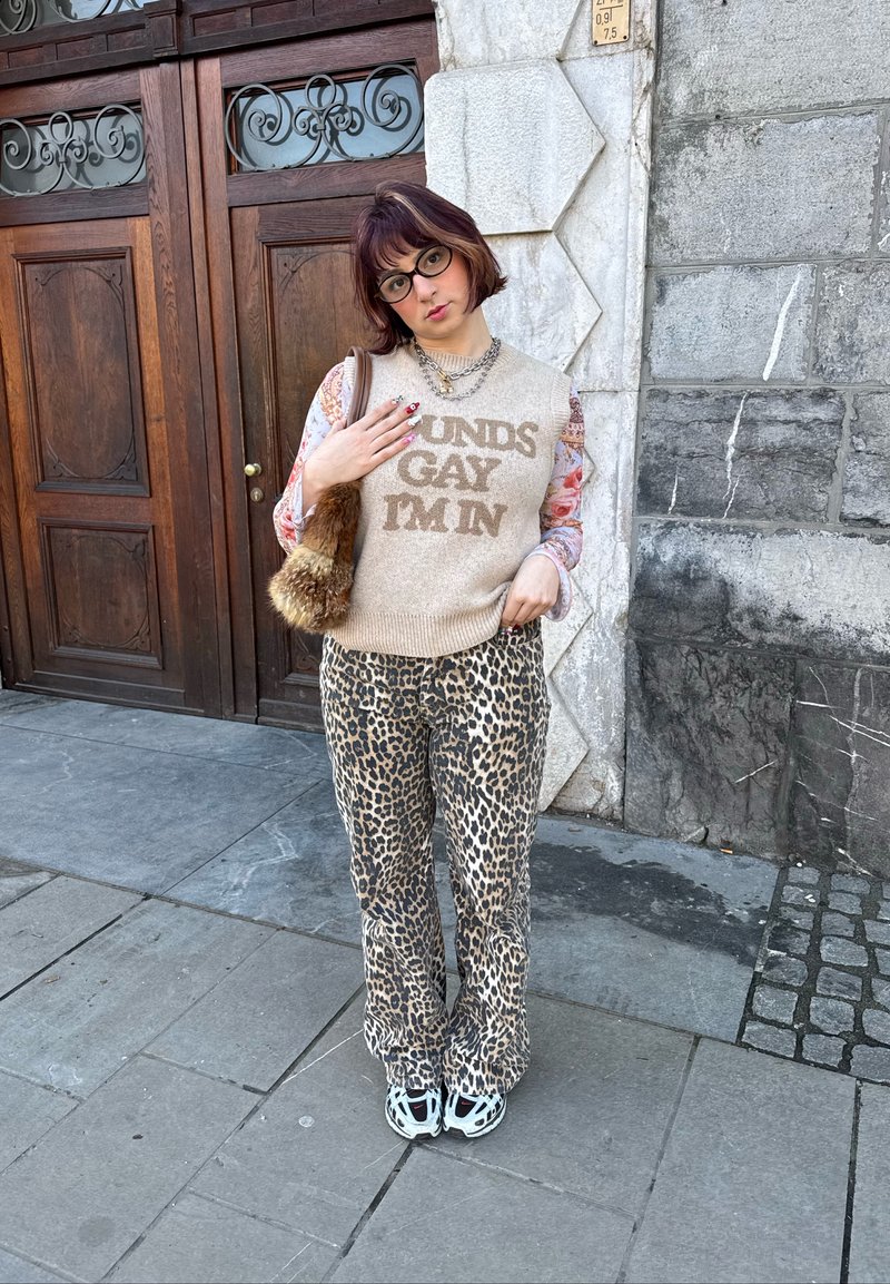 Personne aux cheveux courts et portant des lunettes, vêtue d'un pull beige avec un texte, d'une chemise à fleurs, d'un pantalon à imprimé léopard et de baskets blanches, debout près d'une porte en bois.