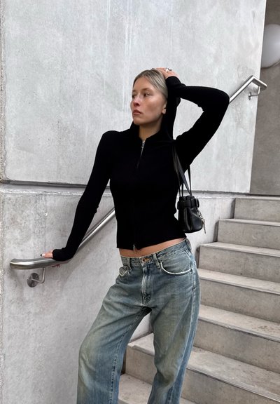 Sudadera con capucha negra de canalé con cremallera, silueta ajustada y capucha, combinada con jeans azul claro de corte holgado y un pequeño bolso negro.