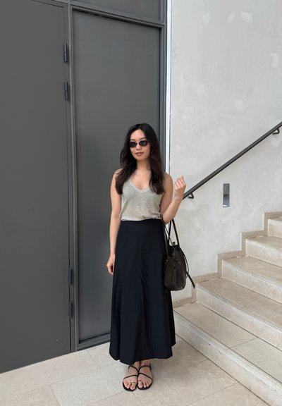 Falda maxi negra, camiseta sin mangas gris con escote en V, gafas de sol negras y sandalias con tiras. Sostiene un bolso oscuro, de pie junto a una escalera de piedra clara.