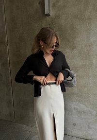 Camisa preta curta com mangas longas, combinada com uma saia branca de cintura alta com uma abertura frontal. Os acessórios incluem um cinto preto e uma bolsa cinza.