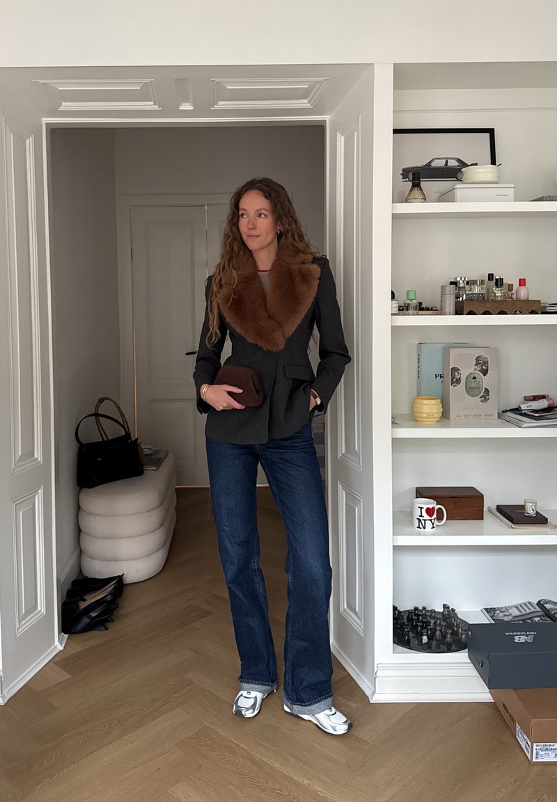 Vrouw met lang krullend haar, gekleed in een donker colbert met een bruine bontkraag, blauwe spijkerbroek en witte sneakers, staand in de deuropening van een moderne kamer.