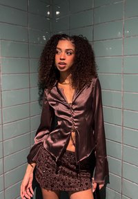 Braunes Satin-Langarmhemd mit Schnürdetail vorne, kombiniert mit einem kurzen, braunen Rock mit Pailletten. Grüne geflieste Wand im Hintergrund.