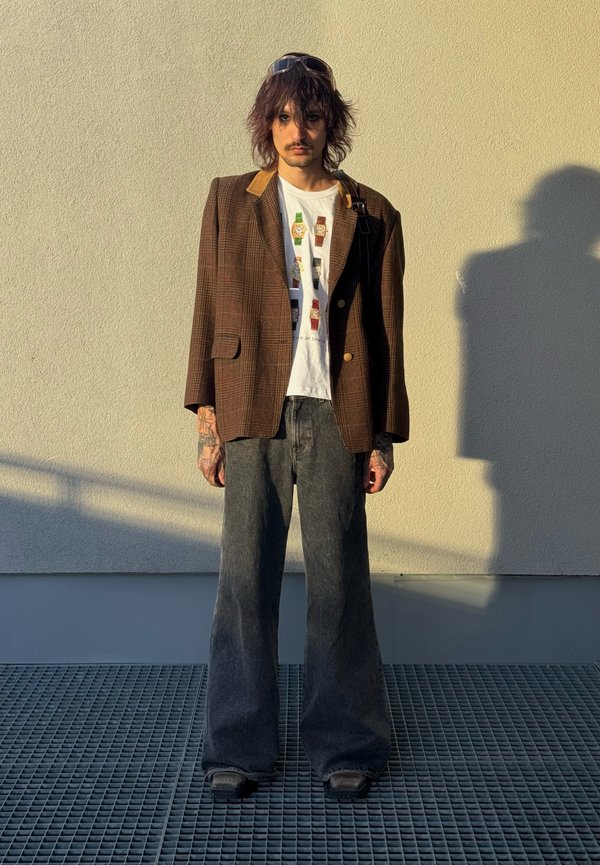 Personne aux cheveux mi-longs portant des lunettes de soleil sur la tête, blazer marron à carreaux, t-shirt blanc graphique, pantalon large gris, debout contre un mur beige.